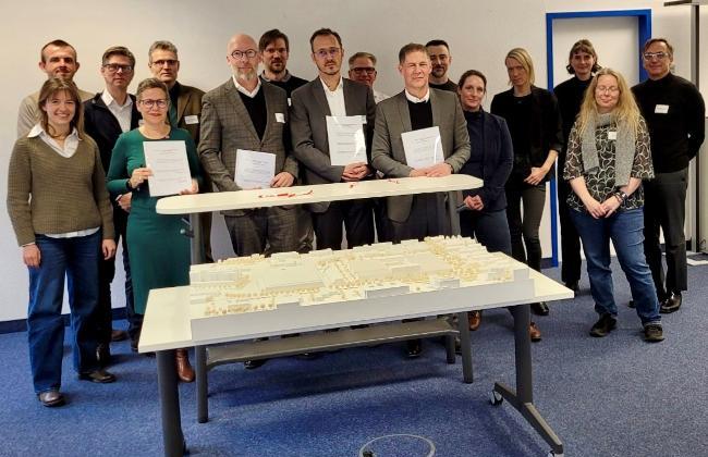 Gruppenbild Projektpartner mit den unterzeichneten Verträgen hinter dem Entwurf des Neubaus Gruppenbild Projektpartner mit den unterzeichneten Verträgen hinter dem Entwurf des Neubaus